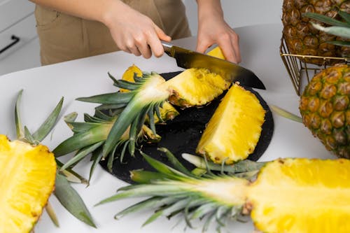 Unrecognizable female cutting ripe pineapples on white table in modern kitchen at home