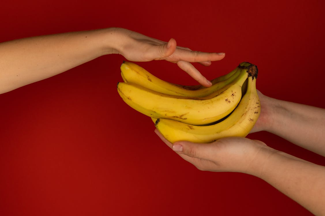 Crop anonymous woman touching smooth yellow peel of fresh banana against partner on red background