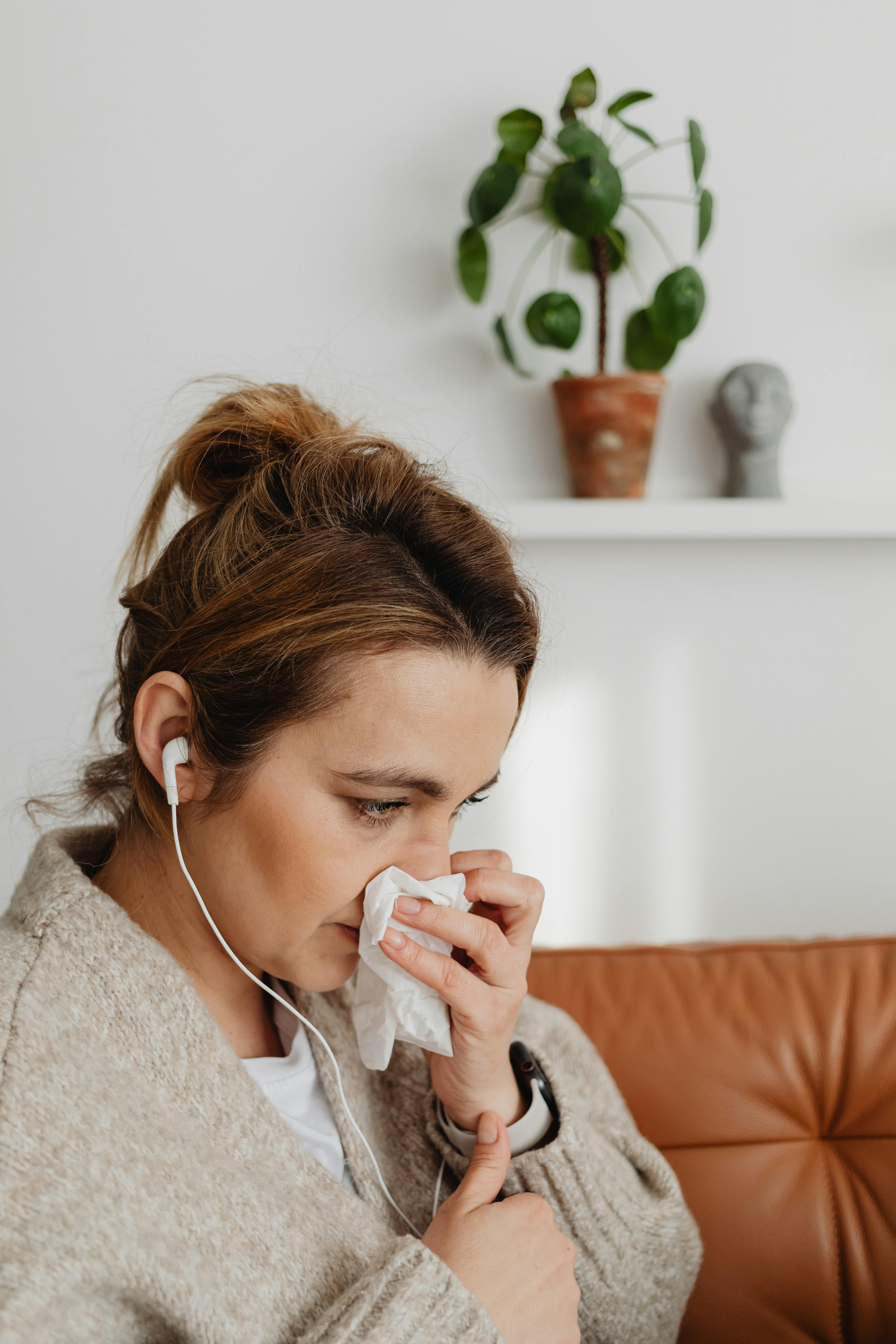 Woman wearing earphones using a tissue, sitting indoors, with a plant in the background.