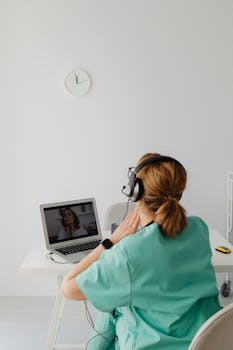 A female doctor in a green uniform conducts an online consultation through a laptop in a bright office.