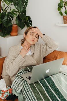 Woman with fever using a laptop while wrapped in a blanket at home, resting on a sofa.