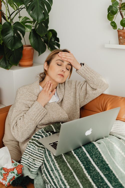 Free Woman with fever using a laptop while wrapped in a blanket at home, resting on a sofa. Stock Photo Free Woman with fever using a laptop while wrapped in a blanket at home, resting on a sofa. Stock Photo