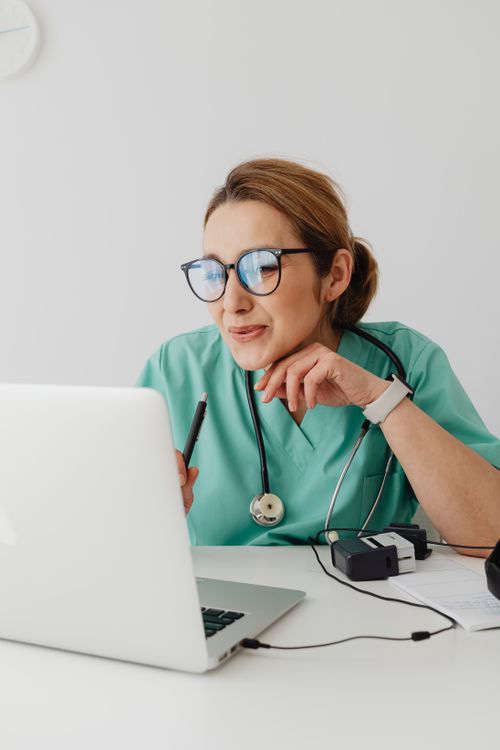 Free A healthcare professional wearing scrubs and eyeglasses using a laptop with a stethoscope around the neck. Stock Photo
