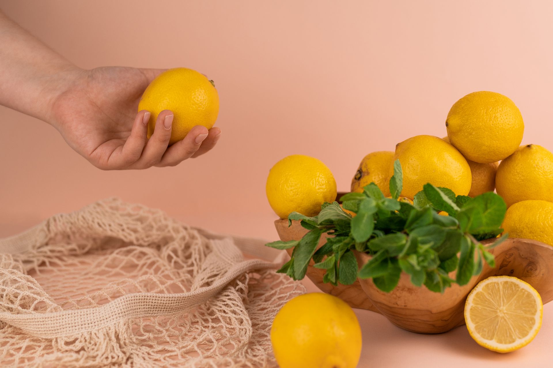 A hand reaches for a fresh lemon next to a bowl of lemons and mint on a pink backdrop.