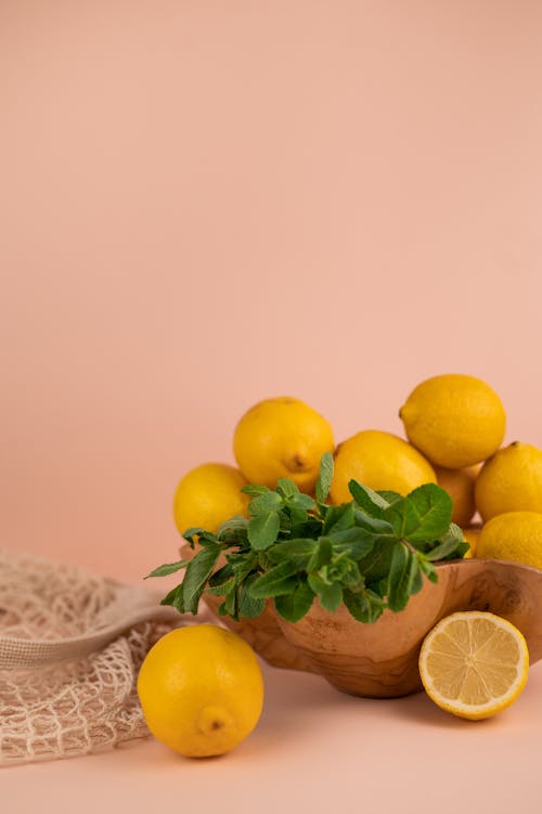 Ripe whole and cut lemons with mint foliage in wooden bowl near zero waste bag on pastel background