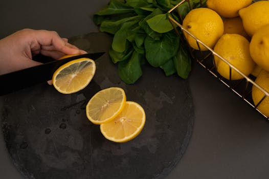 Hand slicing fresh lemons on a dark slate board with spinach and lemons in the background.