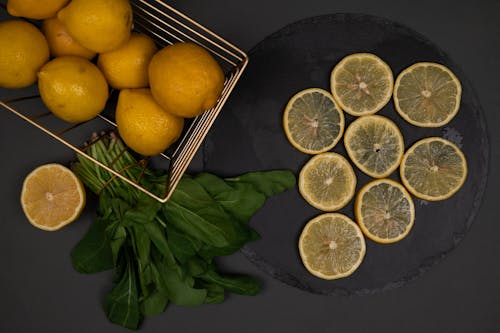 Top view of fresh whole and cut lemons on black chopping board near spinach foliage on dark background