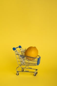 A single lemon sits inside a tiny shopping cart against a vibrant yellow background, creating a minimalist and bright composition.