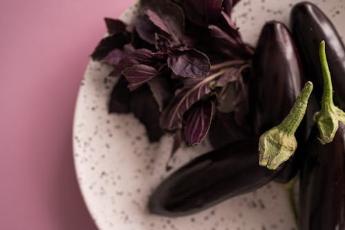 Top view of fresh ripe eggplants with stem and bunch of red basil leaves on plate and on lilac background