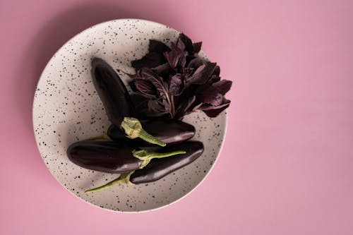 Top view of white speckled plate with fresh dark aubergines and bunch of fragrant red basil herbs on pink surface