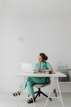 Healthcare professional in scrubs using laptop for telemedicine session in modern office.