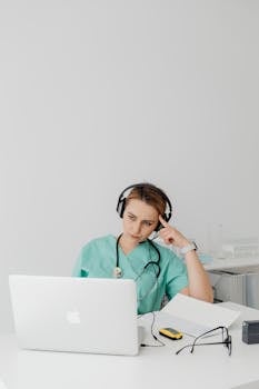 A female doctor in scrub suit using a laptop and headphones for an online video call in a bright office.