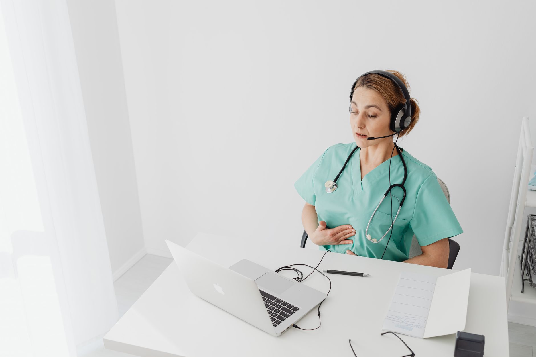 Medical professional using laptop for virtual healthcare session, wearing headset and scrubs.