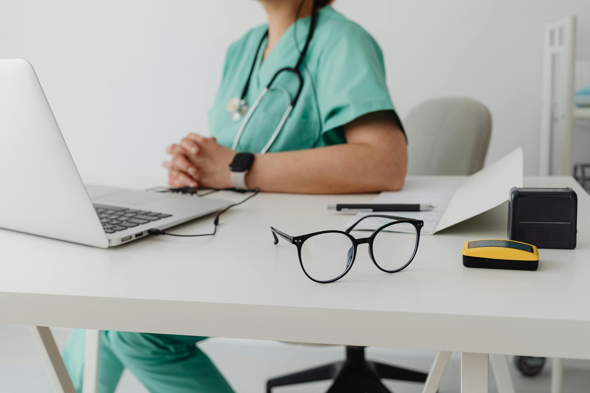 A healthcare professional in scrubs using a laptop in an office setting.