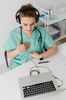 Female doctor in scrubs conducting virtual consultation using laptop and headphones.