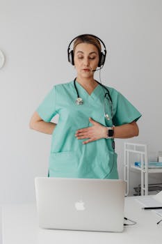 Medical professional in scrubs using a laptop for online consultation with a headset.