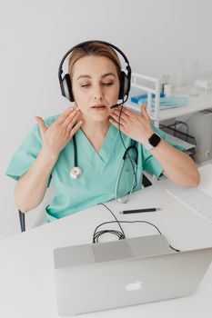 A female doctor in scrubs conducts a virtual consultation using a laptop and headphones.