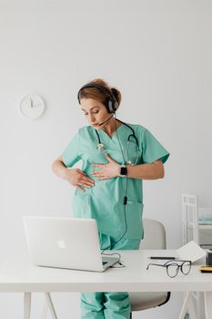 A female doctor in green scrubs conducting a virtual consultation using a laptop and headset, indoors.