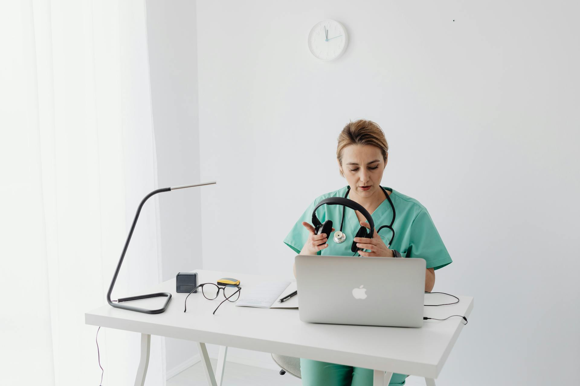 Female doctor in scrubs using a laptop for telehealth consultation in modern office setting.