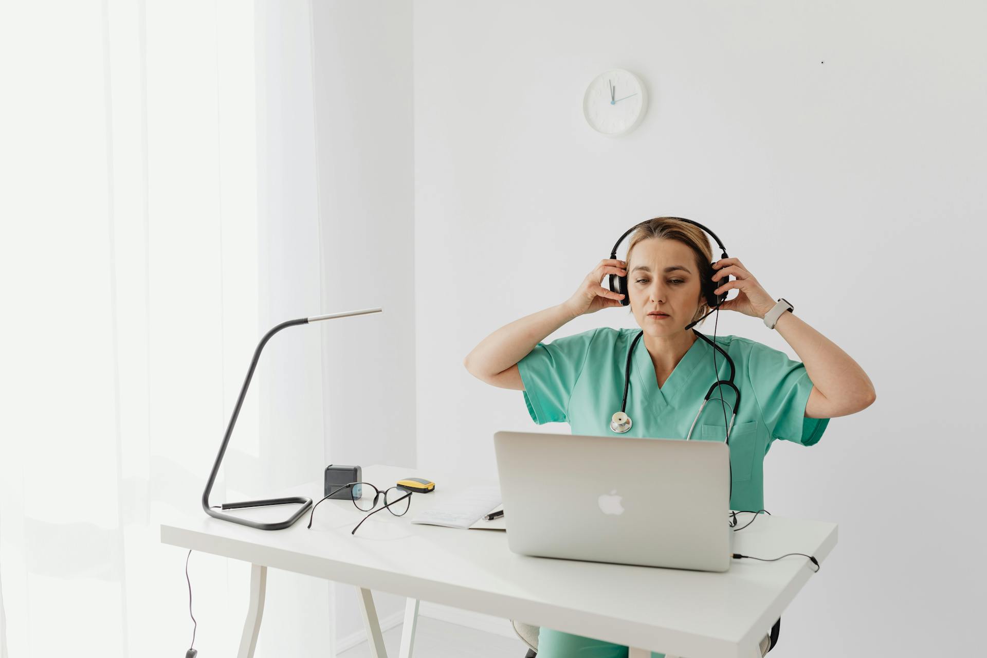 Female healthcare worker uses laptop and headphones at desk in modern office.