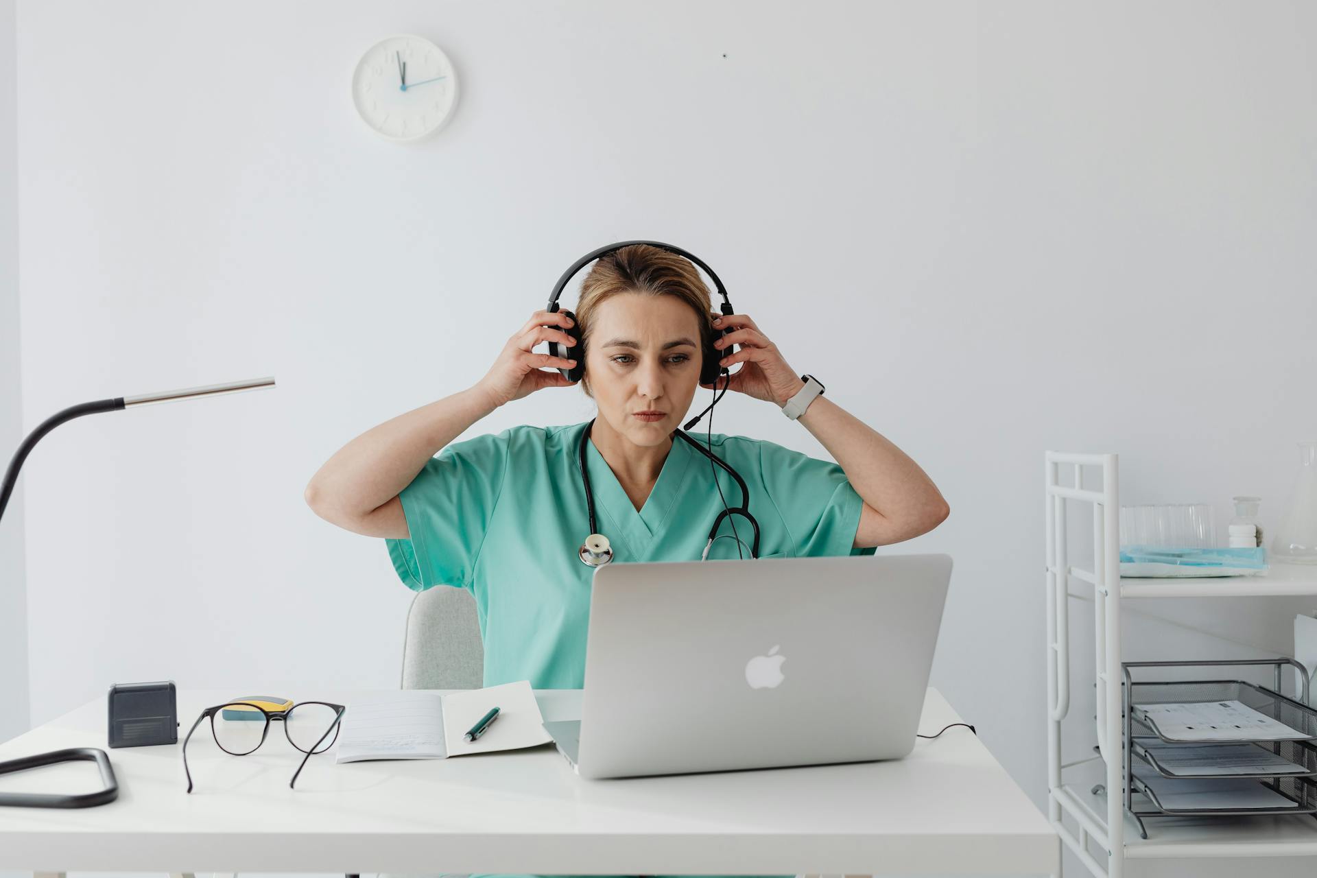 Medical professional in teal scrubs using a laptop and headset, sitting at a desk in an office setting.