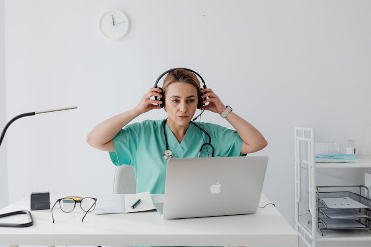 A Woman In Teal Shirt Sitting Near The Table With Laptop While Wearing A Headset