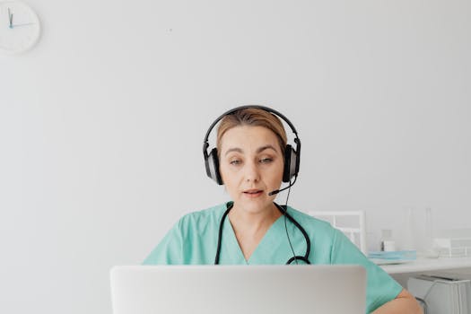 A healthcare worker in scrubs on a video call, focusing intently.