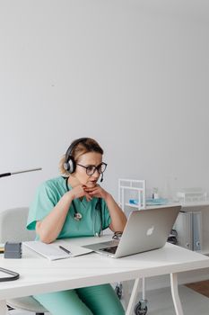 A medical professional focuses intently on a laptop while wearing teal scrubs and headphones.