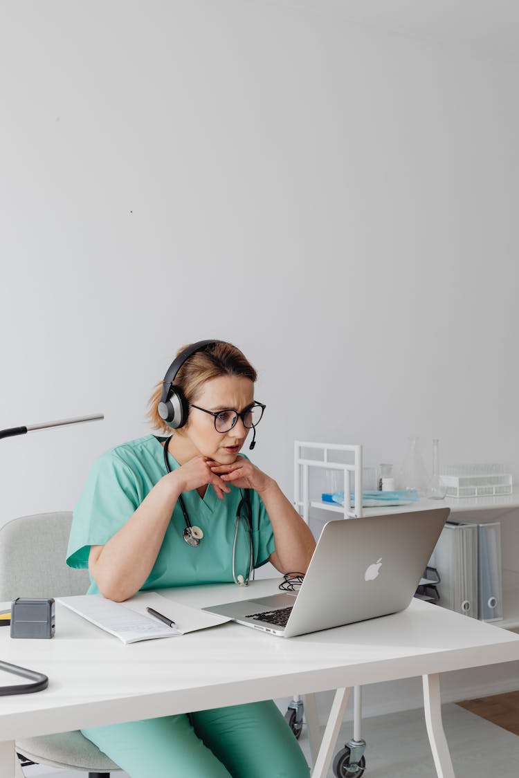 Woman In Teal Uniform Wearing Eyeglasses And Headphones While Looking At The Laptop 