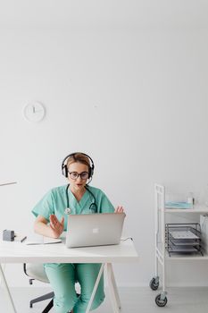 A female doctor in scrubs conducts an online consultation using a laptop in a medical office.