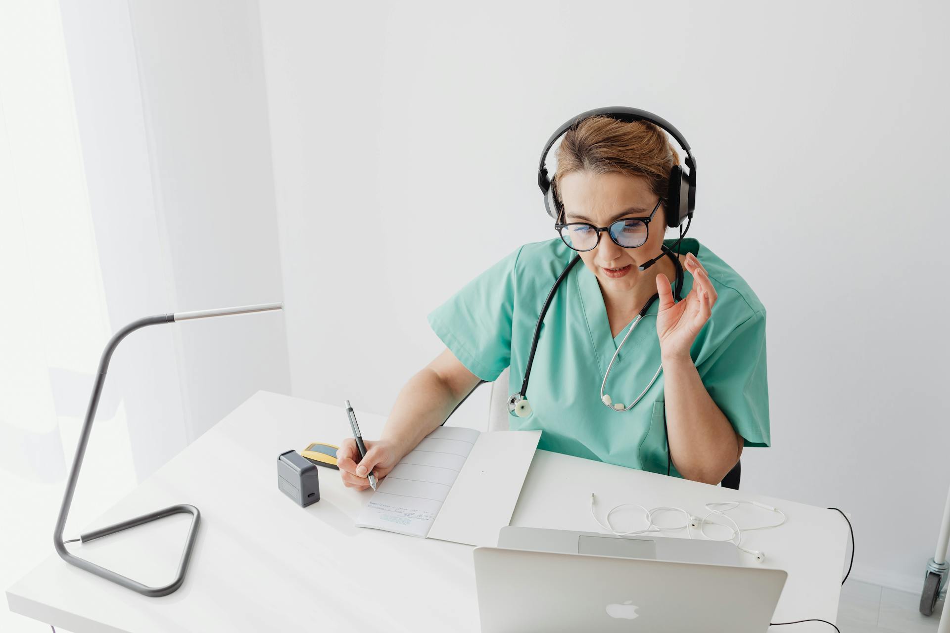 Doctor in scrubs with headset engages in an online consultation while taking notes.