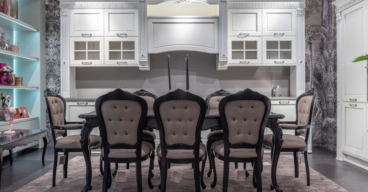 Comfortable chair and big dining table placed in classic styled kitchen with white furniture near illuminated shelves with various decorative items