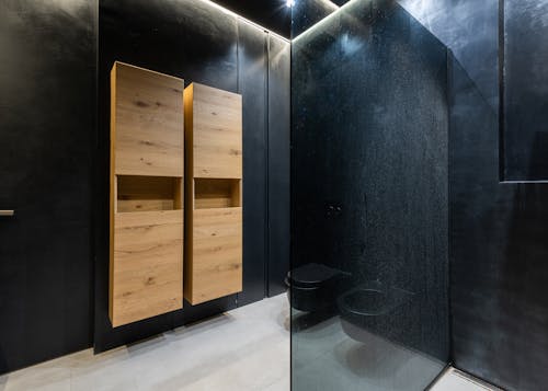 Wooden cupboards hanging on black wall near toilet bowl and shower cabin in contemporary bathroom