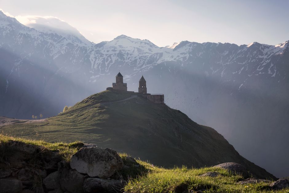 Capturée sous un doux jour, une église se dresse au sommet d'une colline entourée de montagnes et de rayons de soleil.