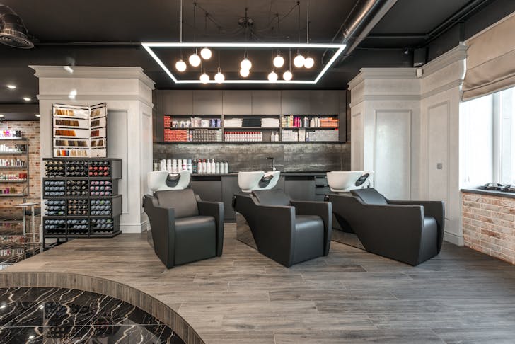 Black leather backwash chairs with sinks under glowing lamps placed in modern spacious beauty salon with cosmetology products and window