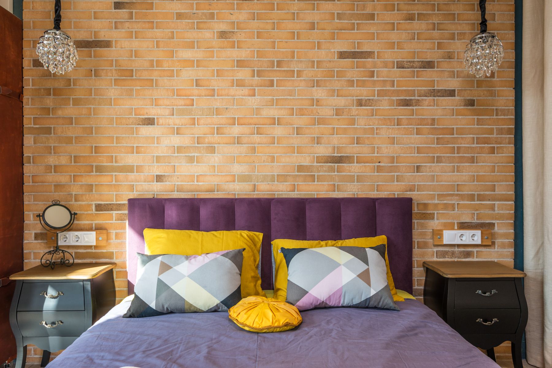 Contemporary bedroom with a purple upholstered headboard, brick wall, geometric bedding, crystal chandelier and warm wood floor