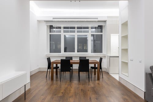 Wooden table with chairs placed near window in light spacious apartment with empty shelves and minimalist interior at home