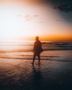 A man stands on the shore as the sun sets over the ocean, creating a serene silhouette.