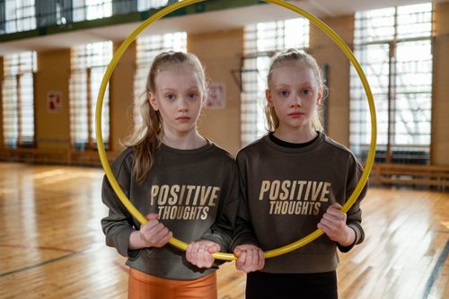 Free Twin girls in gym holding a hula hoop, wearing 'Positive Thoughts' sweatshirts. Stock Photo