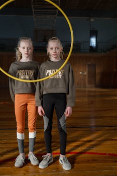 Twin girls stand indoors in a gym with a gymnastics hoop, sporting positive messages.