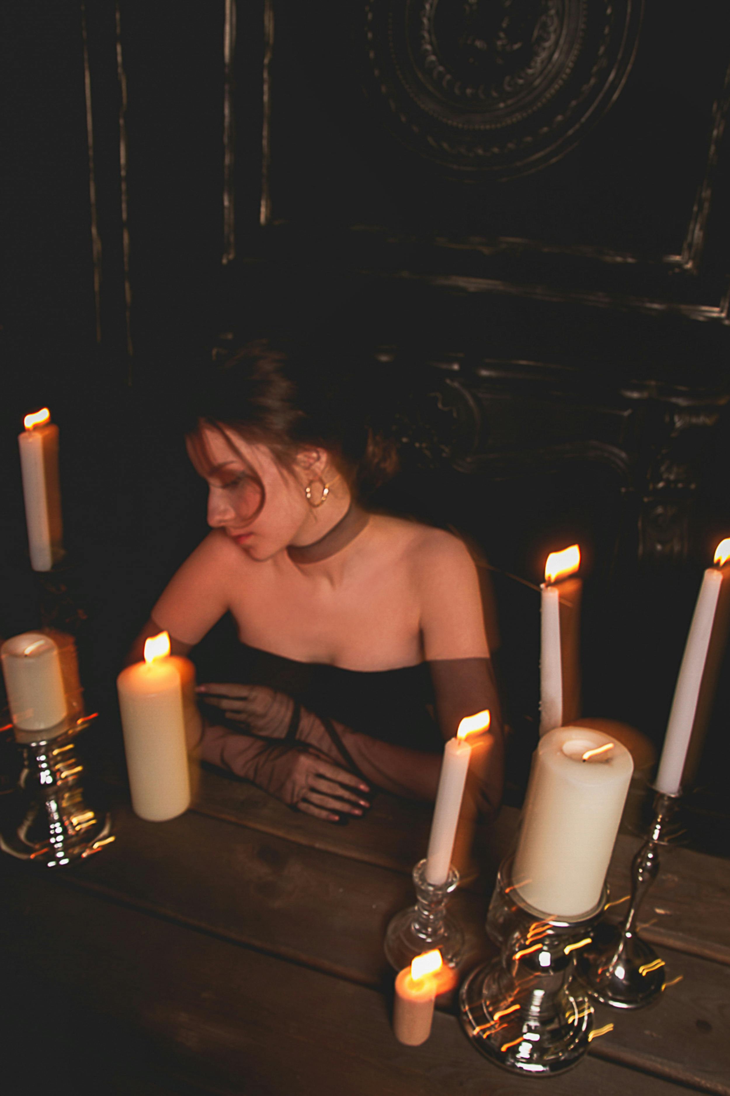 Lady in black clothes sitting at wooden table with various burning wax candles in dark house with ornamental walls