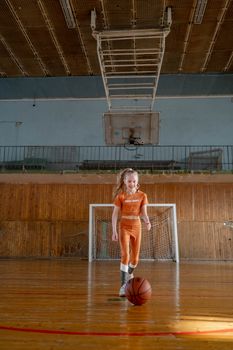 Girl in an orange outfit dribbling a basketball indoors, joyful and active.
