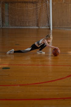 A young girl stretches with a basketball on a wooden indoor court, symbolizing training and exercise.
