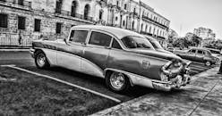 这张黑白照片展示了一辆停在古巴哈瓦那的老爷车，呈现出复古风格和建筑风貌