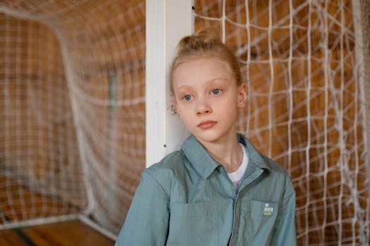 Portrait of a young girl in a green jacket standing by a sports net indoors.