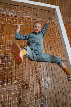 A young girl in a green jumpsuit joyfully hangs on an indoor playground net with a wooden backdrop.