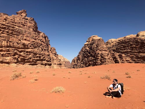 Man Wearing White Shirt and Black Pants Sitting on Soil Behind Rock Formation Mountain