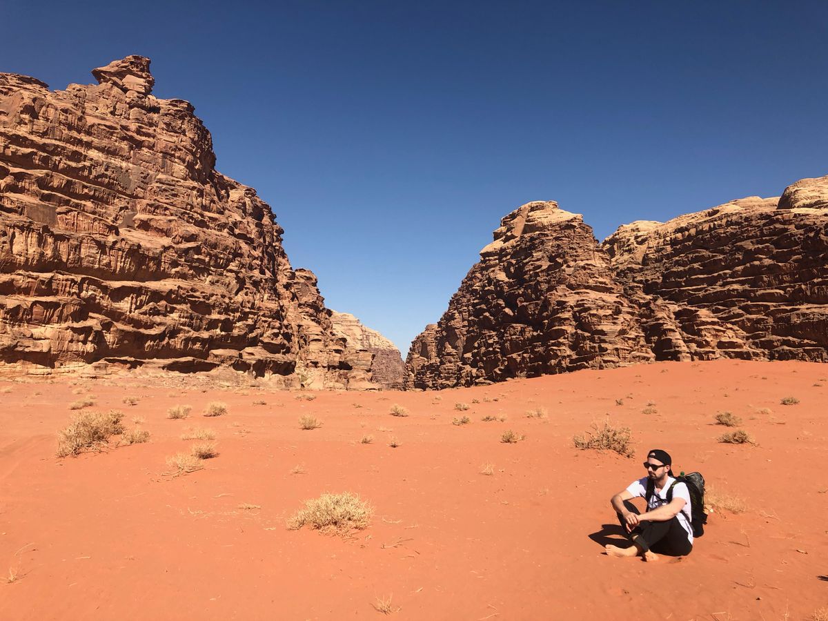 A guide with a backpack sits on red sand at the mouth of a narrow sandstone canyon, tall rock walls rising on either side under a clear blue sky.