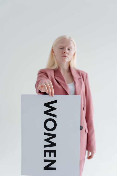 Free A woman in a pink blazer holds a sign with 'Women' text in a bright studio setting. Stock Photo
