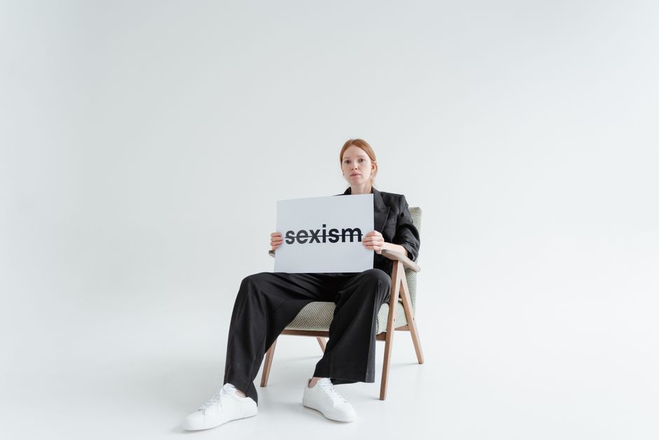 Mujer con traje negro sosteniendo un cartel de 'sexismo', resaltando la igualdad de género en un estudio blanco minimalista.